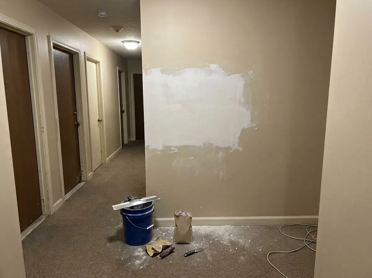 A freshly mudded, unpainted repair on a wall in an apartment building corridor, unit doors along the hall and a bucket, knife and sandpaper on the carpet below