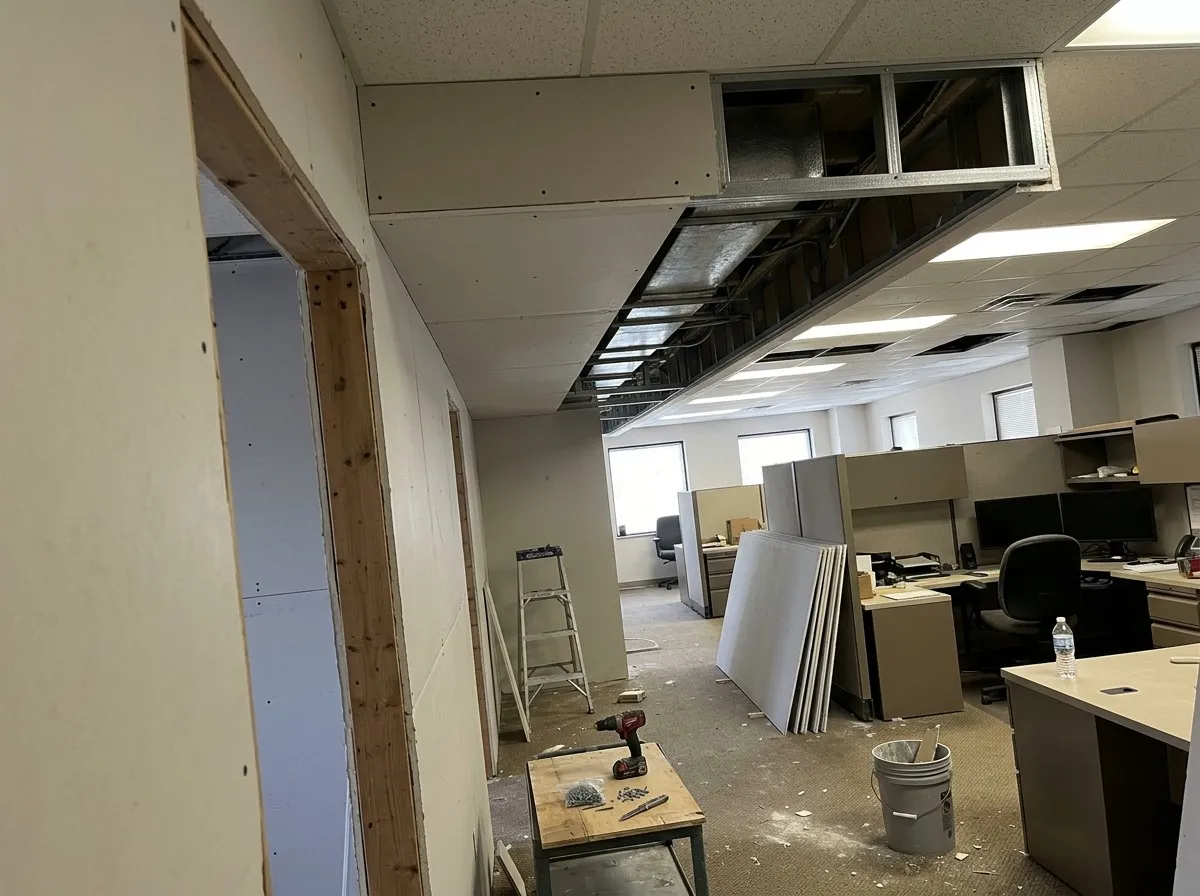 A drywall soffit being built along an office ceiling to box in ductwork, its metal framing still open at one end, beside an acoustic tile ceiling over the cubicles, with sheets of board and tools on the floor
