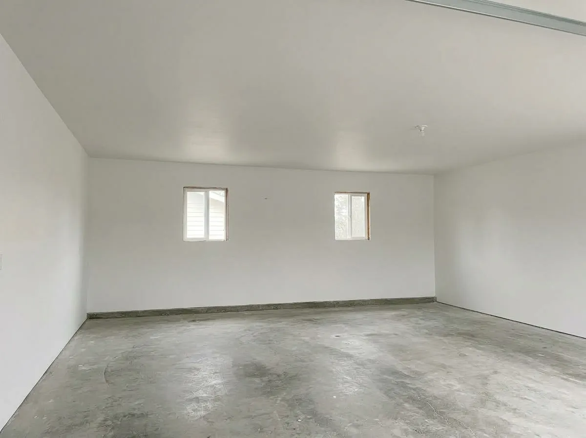 An empty garage with the ceiling and walls finished smooth in drywall, two small windows on the back wall and a bare concrete floor